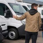Two men discussing a row of white commercial cargo vans at an outdoor vehicle dealership.