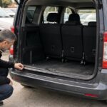 Man in a black hoodie inspecting the open trunk and rear bumper of a grey 5-seater van in a parking lot.